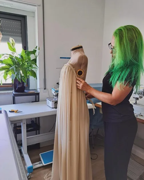 Modista a ajustar à mão um vestido comprido bege no manequim, no atelier JacqueCostura